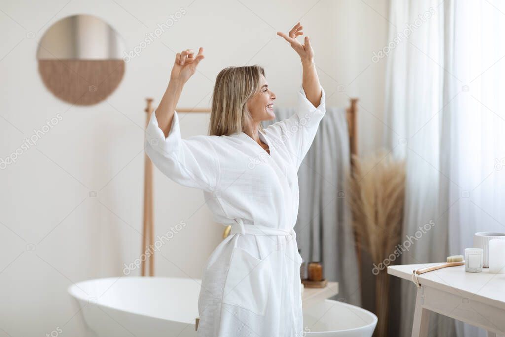 Joyful attractive middle aged woman with blonde hair dancing with hands up in white bathroom at home, wearing long bathrobe, enjoying weekend, going to take relaxing bath, panorama, copy space