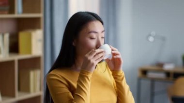 Hoş aile alışkanlıkları. Genç ve güzel Asyalı bir bayanın yakın portresi. Evin içinde kahveden zevk alıyor, gülümsüyor, yan bakıyor, ağır çekim, boş alan.
