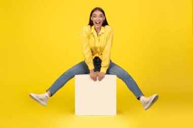 Küpte oturan neşeli milenyum kadını sarı arka planda kameraya bakıp gülümsüyor. Carefere Female stüdyoda eğleniyor. Tam Uzunluk Çekimi