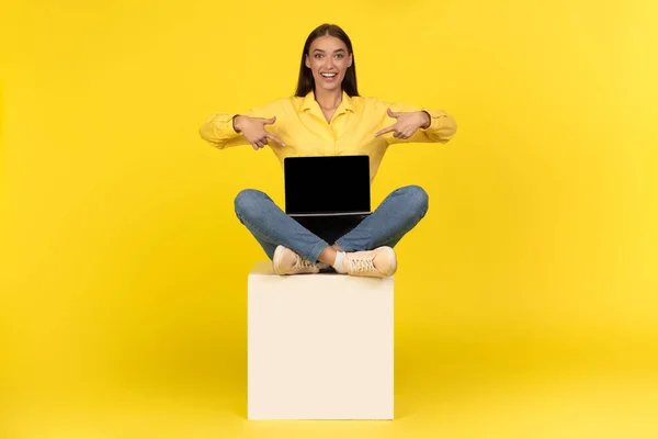 Cheerful Lady Pointing Fingers At Laptop With Blank Screen Advertising Website Or Internet Service Sitting On Cube Over Yellow Studio Background. Female Freelancer Showing Computer. Mockup