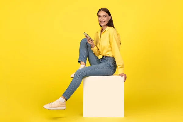 Cheerful Lady Using Cellphone Smiling Camera Sitting Cube Yellow Studio — ストック写真