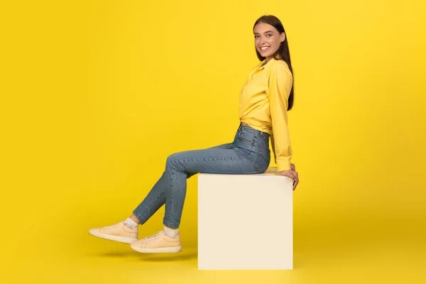 Cheerful Millennial Woman Smiling To Camera Sitting On Cube Posing Over Yellow Background. Studio Shot Of Happy Female. Beauty And Style. Full Length Shot