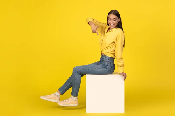 Cheerful Lady Sitting Cube Pointing Finger Posing Smiling Camera Yellow — Stock Photo, Image