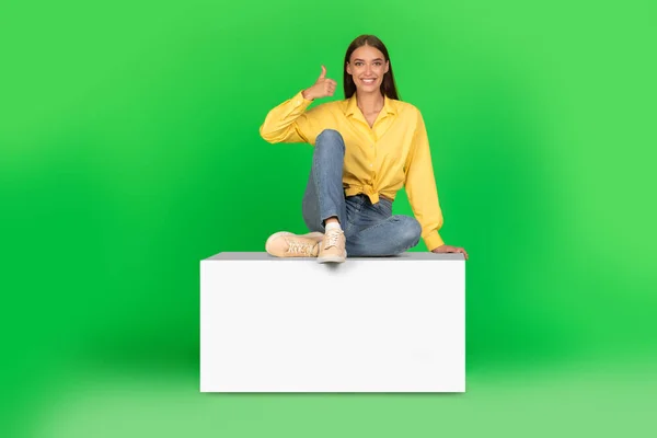 Cheerful Woman Gesturing Thumbs Smiling Camera Sitting Cube Green Studio — Zdjęcie stockowe