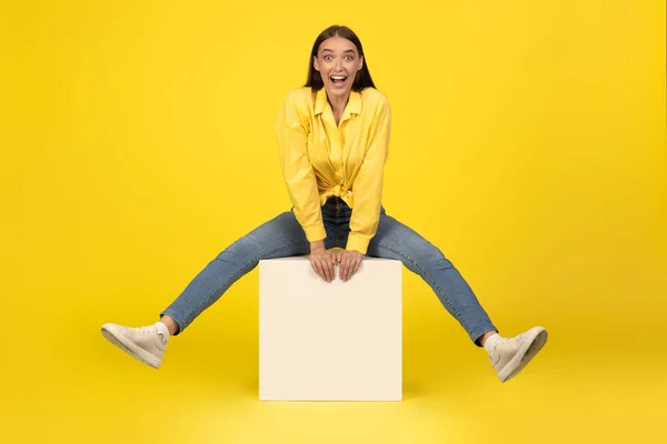 Joyful Millennial Woman Sitting On Cube Smiling Looking At Camera Over Yellow Background. Carefere Female Having Fun Posing In Studio. Full Length Shot