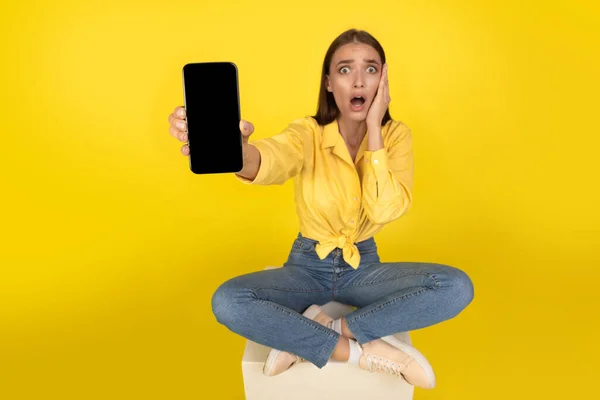 Shocked Woman Showing Smartphone Blank Screen Touching Face Sitting Yellow — Stock Fotó