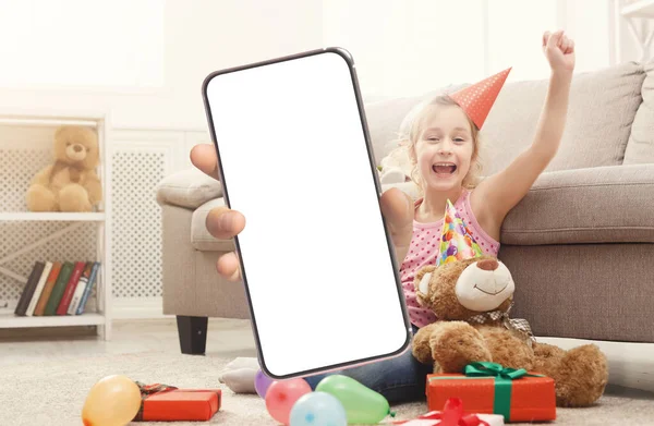 Overjoyed Little Girl In Birthday Hat Holding Big Blank Smartphone, Excited Cute Female Child Showing Mobile Phone With White Screen And Exclaiming With Joy, Recommending New App, Mockup