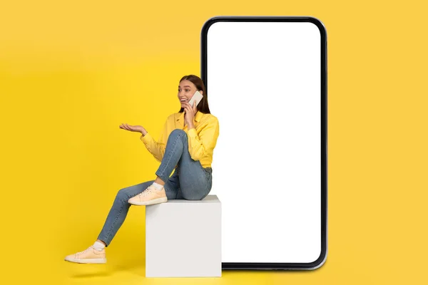Excited Woman Talking On Cellphone Sitting On Cube Near Huge Smartphone With Blank Screen Over Yellow Studio Background. Mobile Communication. Mockup, Collage