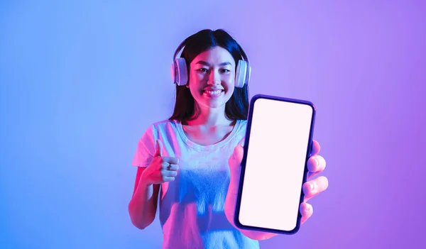 Music App. Happy Young Asian Woman Wearing Headphones Showing Big Blank Smartphone With White Screen While Standing Under Neon Light, Smiling Korean Female Gesturing Thumb Up, Collage, Mockup
