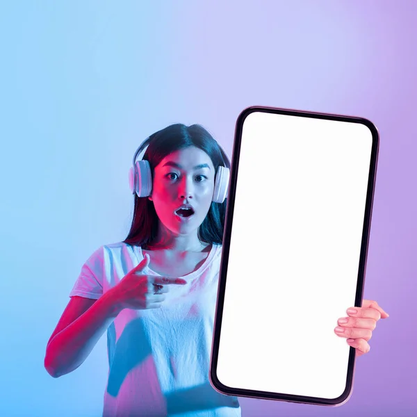 Cool App Shocked Asian Female Headphones Pointing Big Blank Smartphone — Stockfoto