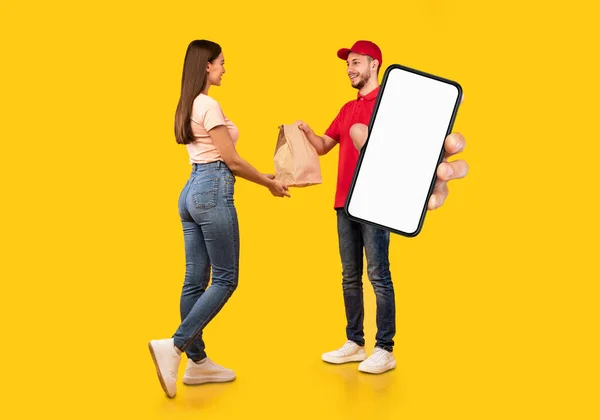 Delivery App. Male Courier Holding Big Blank Smartphone And Giving Package To Female Customer, Recommending Application Or Website, Standing Together Over Yellow Background In Studio, Mockup, Collage