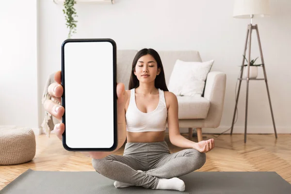 Yoga App. Calm young asian woman meditating and holding big blank smartphone in hand, relaxed female sitting in lotus position with closed eyes, demonstrating copy space for design, collage, mockup