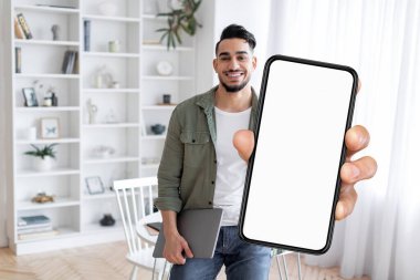Online Ad. Smiling Arab Man Demonstrating Smartphone With Big Blank Screen While Standing Near Desk At Home, Happy Middle Eastern Guy Recommending New Mobile App Or Website, Collage, Mockup