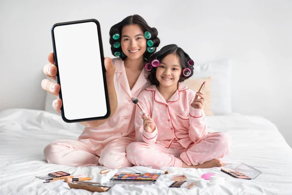 Smiling Asian Mother And Daughter In Pajamas Showing Blank Smartphone At Camera, Korean Mom And Female Child Recommending Beauty App While Sitting On Bed And Doing Makeup Together, Collage, Mockup