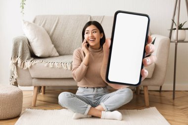 Mutlu Asyalı kadın telefonda konuşuyor ve evde otururken kamerada büyük boş akıllı telefonu gösteriyor. Neşeli kadın telefon görüşmesi, kolaj, taklit, yeni uygulama öneriyor.