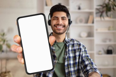 Mobile Advertisement. Cheerful Arab Guy In Headset Holding Big Blank Smartphone With White Screen While Sitting On Couch At Home, Middle Eastern Man Advertising New App Or Website, Collage, Mockup