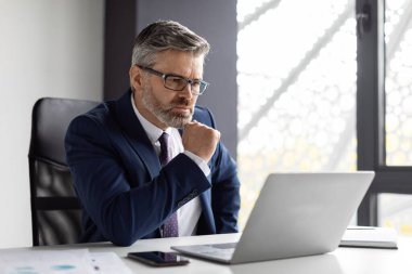 Ofiste çalışırken dizüstü bilgisayara bakan odaklanmış orta yaşlı işadamı, masada oturan ve bilgisayar kullanan, şirket e-postalarını okuyan, serbest alan okuyan, iş adamı, yetişkin erkek girişimci.