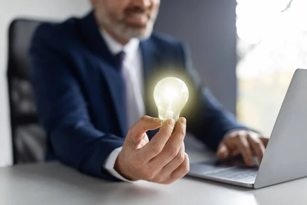 Business Innovations. Middle Aged Businessman Holding Illuminated Light Bulb While Sitting At Workplace With Laptop Computer, Entrepreneur In Suit Having Startup Idea, Closeup Shot, Cropped