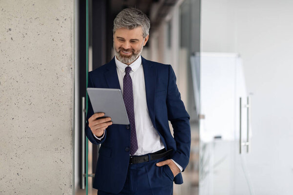 Handsome middle aged businessman using digital tablet while standing in office building corridor, smiling male entrepreneur in suit holding modern gadget, browsing app for business, copy space