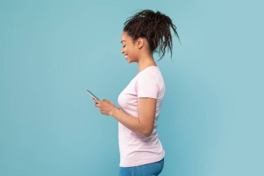 Akıllı telefon kullanan siyahi bir kadının profil portresi, SMS mesajı yazmak, arkadaşla sohbet etmek, mavi stüdyo arka planında internet gezmek, kopyalama alanı, kenar görünüm
