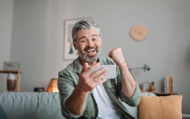 Sakallı tatmin olmuş yaşlı Avrupalı adam akıllı telefona bakıyor ve elleriyle başarıya işaret ediyor ve odanın içindeki zafere seviniyor. Çevrimiçi kazanç, oyun ve insanların duyguları, harika bir teklif ve yeni uygulama