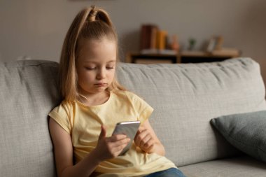 Sosyal medyada cep telefonu şebekesini kullanan mutsuz küçük kız evdeki koltukta oturuyor. Sıkıcı çocuk cep telefonuyla oyun oynuyor. Mobil Uygulama, Aletler ve Çocuklar Konsepti
