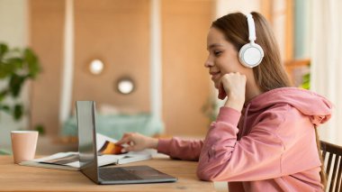 Distance education. Female teenager in wireless headphones studying online on laptop at home, side view, panorama with free space. Schoolgirl using laptop to make homework assignment