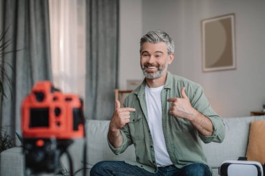 Cheerful old caucasian man blogger with beard records video review, look at camera and points fingers at himself in room interior. Hobby at home in free time, modern work remote, advice to follover