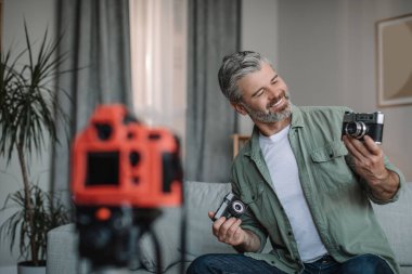 Happy senior european male blogger with beard records video review on photo cameras in room interior, copy space. Comparison of devices, hobby at home in spare time, modern work due social distance