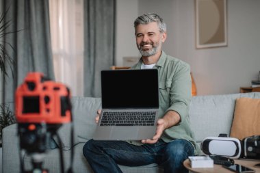 Happy friendly senior european man blogger with beard records video review on technology, shows laptop with empty screen in room interior. Modern work, hobby and vlog on devices at home in free time