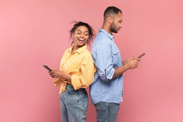 Excited young black lady looking at her boyfriends smartphone. African american couple using cellphones, standing isolated over pink studio background