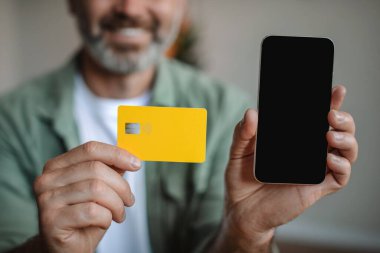 Smiling elderly european man with beard shows smartphone with blank screen and credit card in room, cropped. Sale, online shopping, application for purchases, finance and banking with new device