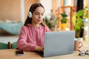 Positive schoolgirl in headphones watching video lesson remotely on laptop, sitting at desk at home interior. Schooling, modern education and knowledge in internet