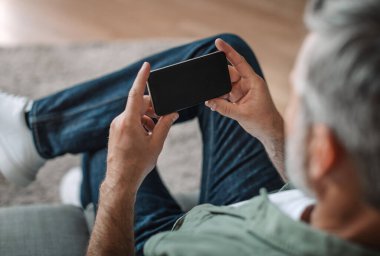 Elderly european man with beard plays in online game or watches video on smartphone with blank screen in room interior. Device for pensioner at home, new app and chat in social networks, ad and offer