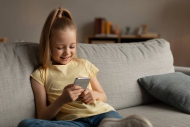 Smartphone kullanan mutlu küçük kız internette oyun oynuyor evinde koltukta otururken eğleniyor. Cep telefonunda mesajlaşan çocuk. Harika Uygulama, Çocuk ve Gadget Konsepti