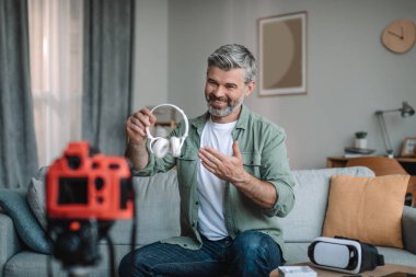 Smiling senior caucasian bearded man blogger records video review on camera, shows headphones, make review in room interior, copy space. Work for retired at home, hobby and overview of devices remote