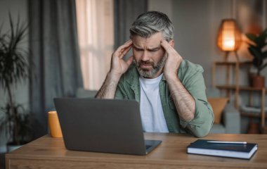 Sad elderly european man with beard looks at laptop at workplace, suffers from headache in living room interior. Brainstorm, migraine from overwork, difficult at work, health problems and deadline
