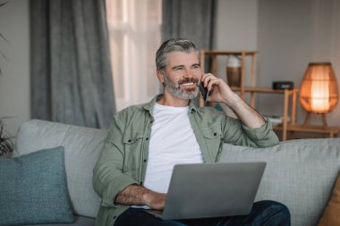 Sakallı neşeli yaşlı Avrupalı adam müşterisini telefonla arıyor ve oturma odasında dizüstü bilgisayarla çalışıyor. Toplantı ve modern teknoloji, iş kumandası, sosyal uzaklık ve evde serbest çalışma