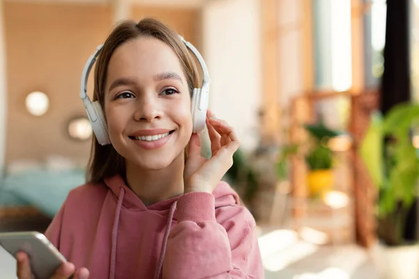 Excited girl in wireless headphones listening music on smartphone, enjoying new audio book, sitting at home, communicating online and smiling, free space