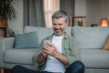 Happy elderly european man with beard typing on smartphone has chat in social media in living room interior. Modern device with new application, blog at home and social distance, good ad and offer