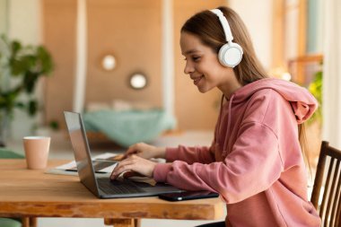 Web tabanlı eğitim konsepti. Kulaklıklı mutlu genç kız evde dizüstü bilgisayar dersi alıyor. Webinar 'a katılan pozitif genç kız, bilgisayar konferansına katılıyor.