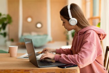 Smart teen girl wearing headphones, having online lesson on laptop, sitting at desk at home, side view, free space. Teenager participating in webinar, learning remotely indoors