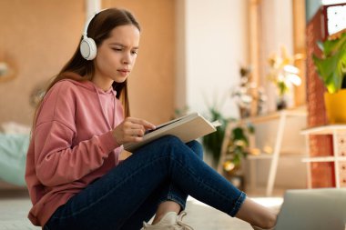 Smart focused schoolgirl with educational materials, books and laptop working on school project at home. Teen girl studying online, writing essay or doing homework