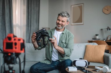 Smiling aged european man blogger look at camera, talk and records video review on vr glasses, enjoy shooting vlog in living room interior, copy space. Entertainment, hobby at home and video call