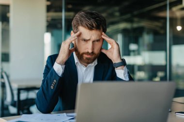 Depresyondaki orta yaşlı işadamı dizüstü bilgisayara bakıyor ve kafasına dokunuyor, baş ağrısı çekiyor ya da sorun yaşıyor, işyerinde oturuyor, boş yer var.