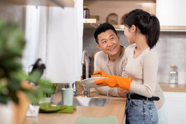 Karısı bulaşık yıkarken, konuşurken, gülümserken, lastik eldiven takan bir kadın, fotokopi çekerken, mutlu koca, orta yaşlı Asyalı bir adam karısını destekliyor.