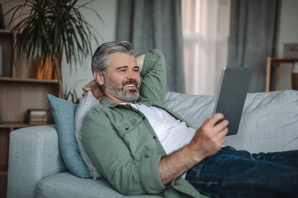 Cheerful old caucasian man resting on sofa and watching video on tablet in living room interior, copy space. Device for pensioner at home, new app for relax, chat in social networks in free time