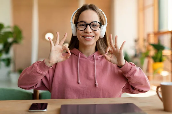Web-based education concept. Portrait of happy girl wearing wireless headphones studying at online school and geturing ok sign, webcam view