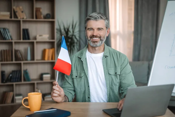 Glad senior caucasian man teacher with beard hold flag of France near blackboard in room interior. Education and online lesson of new language remotely, study at home and international communication
