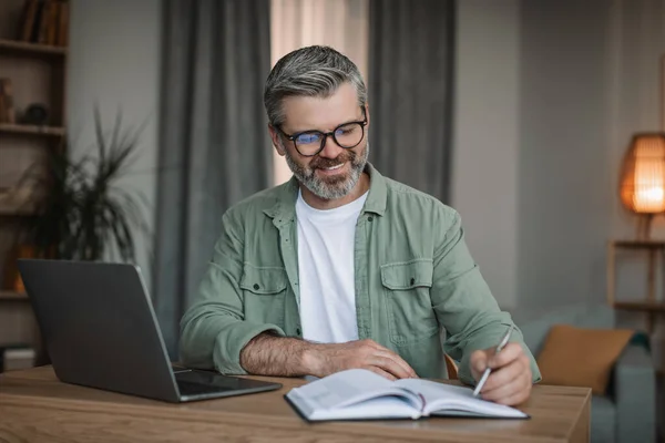 Smiling elderly european male with beard working with laptop, watch video lesson or webinar in living room interior. Modern device and teaching at home, study and social distancing, business remotely